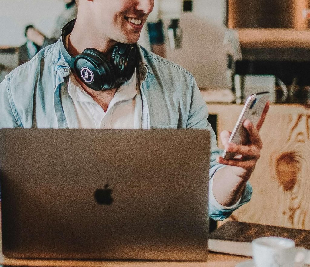 man in coffee shop | telehealth therapy in colorado | telehealth in englewood, co | online therapy in colorado | telehealth in littleton, co | online therapy for college students | emdr therapy colorado | 80125 | 80121 | 80110 Image of a young man, sitting in a coffee shop at a table, smiling, while wearing headphones around his neck and holding a phone. Discover how EMDR therapy can help you cope with past trauma with telehealth in Colorado.