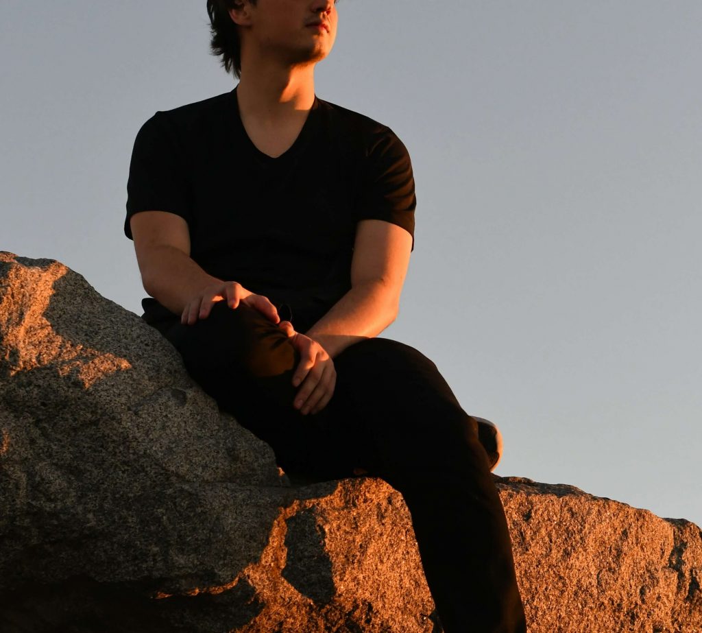 man on rock | neurofeedback therapy for denver, co | neurofeedback therapist in denver, co | neurofeedback for anxiety | neurofeedback for adhd | neurofeedback near me | 80127 | 80128 | 80124 Image of a young man wearing all black sitting on a rock while the sun shines on him as it sets. Learn to manage your ADHD symptoms with the help of neurofeedback therapy in Denver, CO.